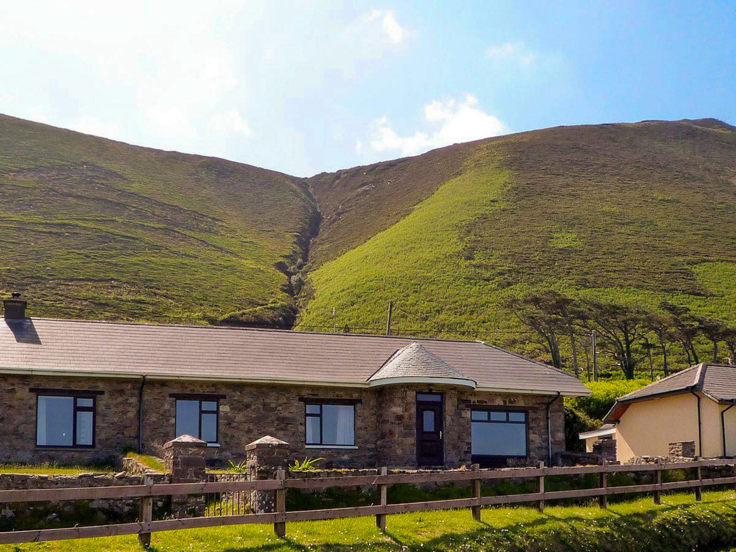 A stone building with a flat roof, a fenced yard, and a valley with hills in the background.
