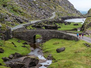 A stone bridge over a stream in a rural landscape with rocky terrain and a body of water in the background. - The Lodge Rossbeigh (Glenbeigh)