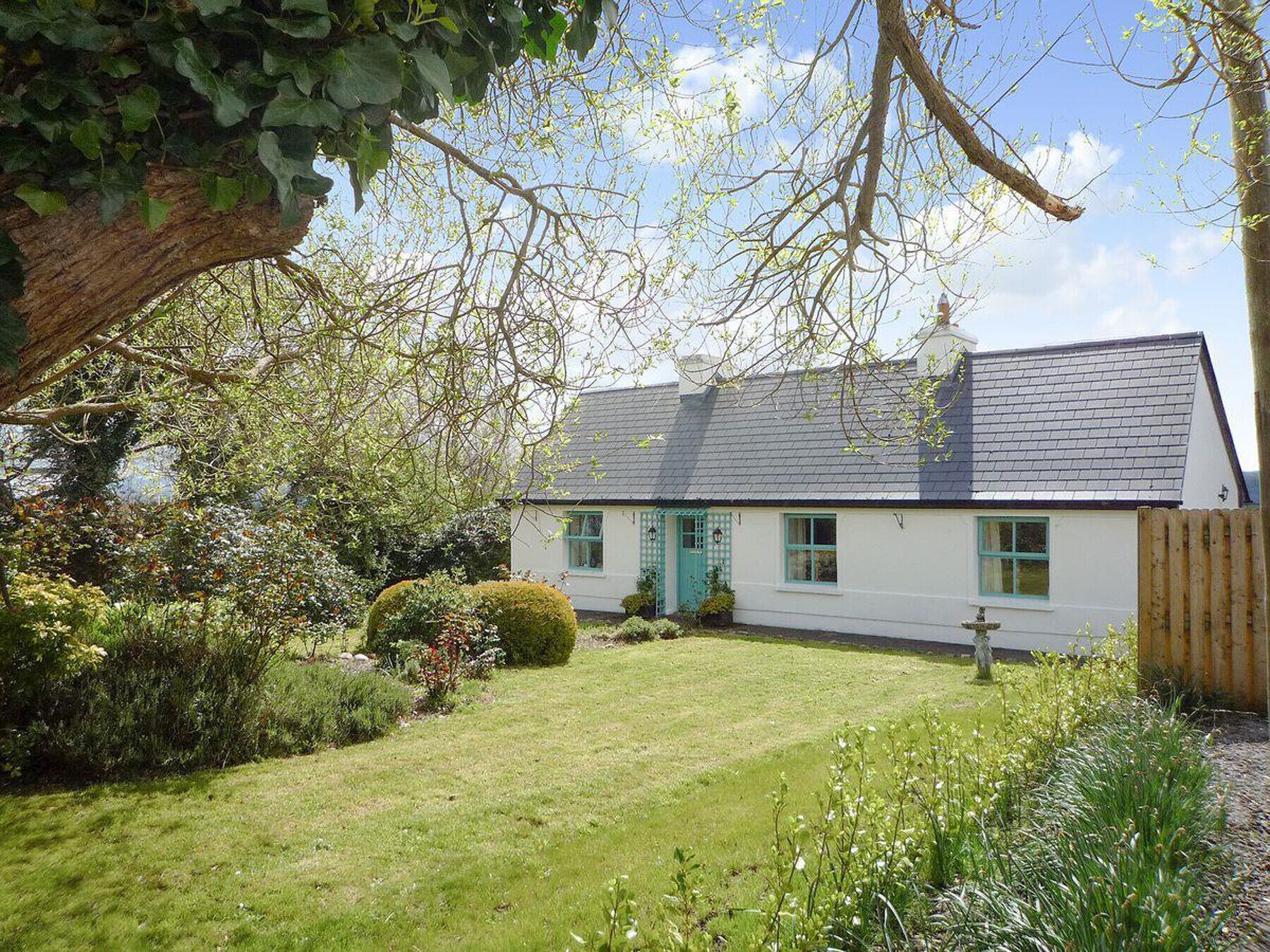 A white cottage with a grey roof, surrounded by a well-maintained garden and a large tree.