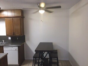 A compact kitchen with wooden cabinets, a dishwasher, a dining table with two chairs, and a ceiling fan.