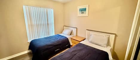 Quarto com duas camas, uma janela com persianas, uma mesa de cabeceira e uma luminária no teto.