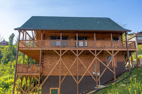 A two-story log cabin with a balcony on the upper floor, surrounded by greenery. - A+ Amazing Views (Gatlinburg)