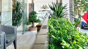 Una entrada de hotel con un diseño moderno, que incluye fachada de vidrio, un cartel con la leyenda 'Queen Ann Hotel' y un camino bordeado de follaje.