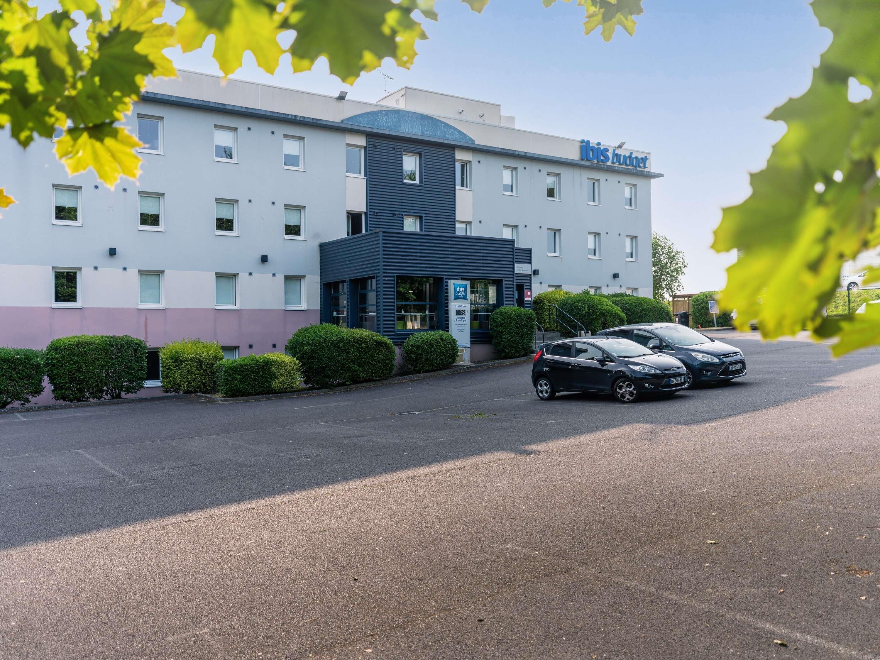 Un hotel de diseño moderno con estacionamiento y varios coches, una entrada destacada y un cartel que dice 'ibis budget'.