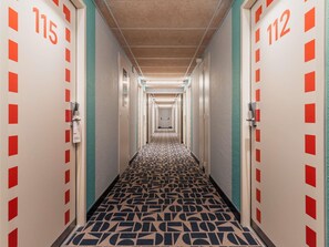 Un pasillo de hotel con puertas numeradas 115 y 112, alfombrado con un diseño geométrico, y un techo con iluminación empotrada.