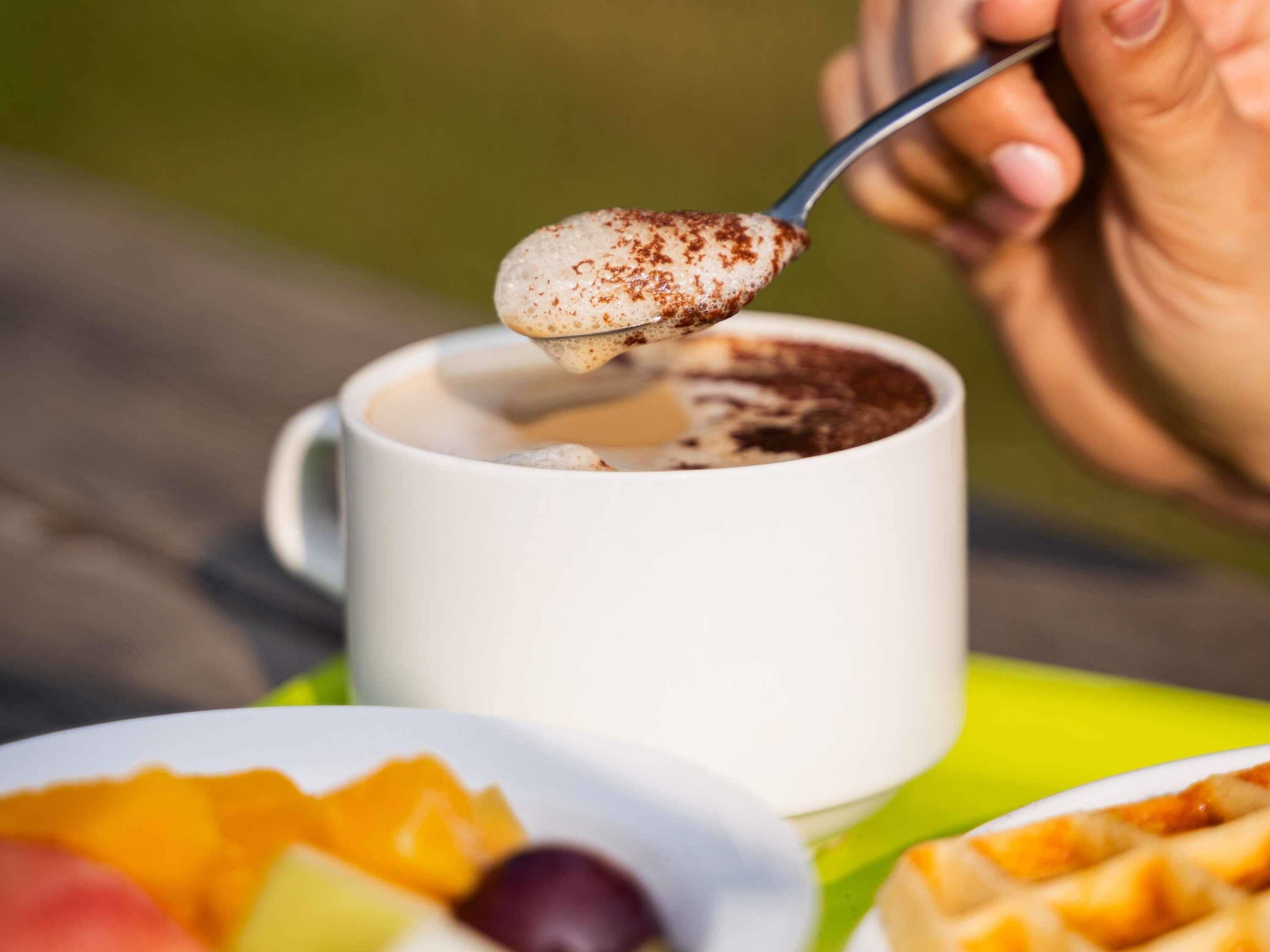 Una mano tiene un cucchiaio sopra una tazza di caffè con schiuma, un piatto di frutta e uno waffle su una superficie verde.
