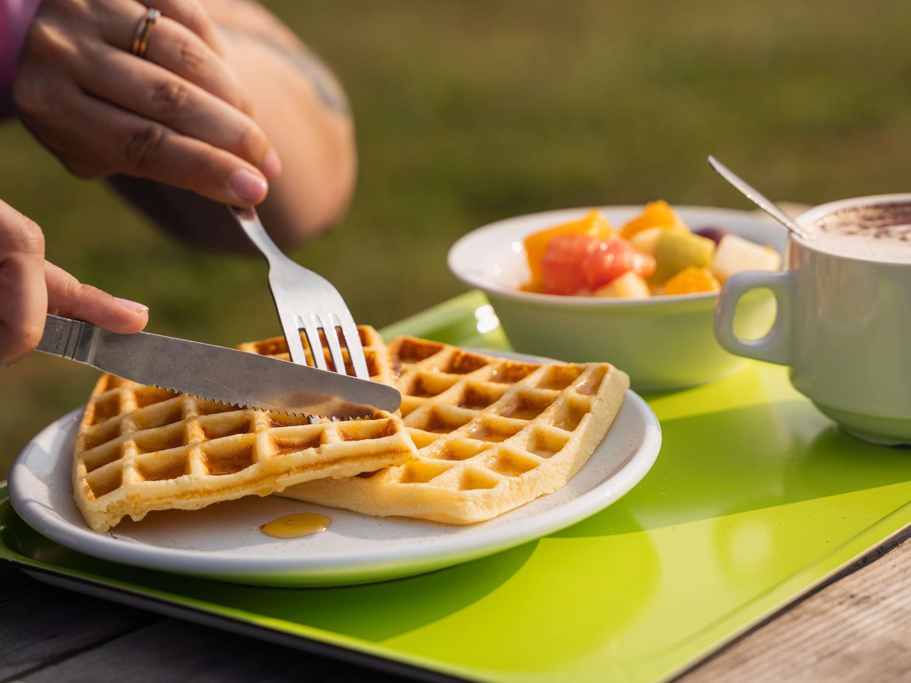 Un gofre en un plato con almíbar, una taza de café y un bol de fruta.