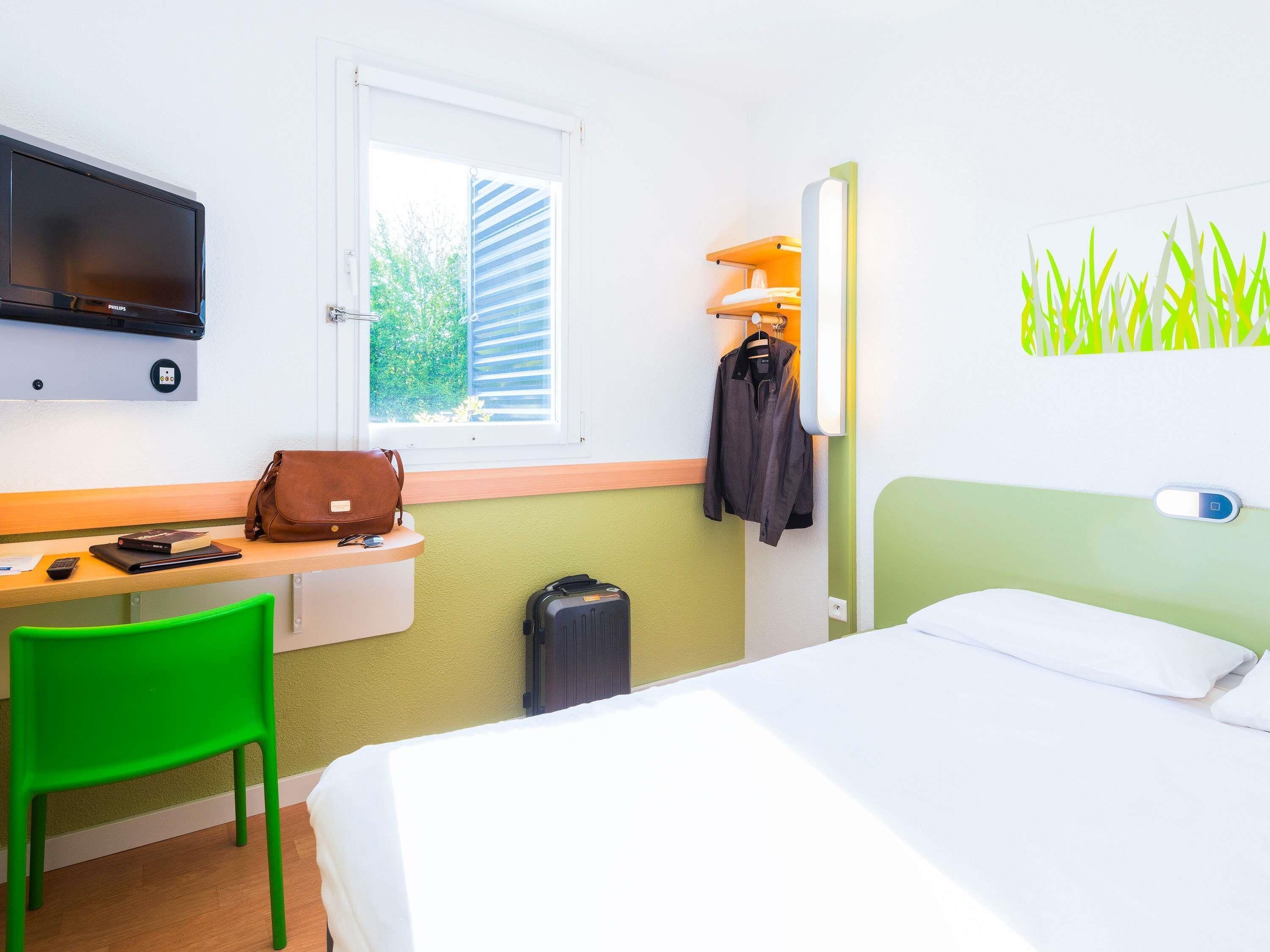 A hotel room with a bed, a desk with a chair, a television, a window with blinds, and a wall with a green plant.