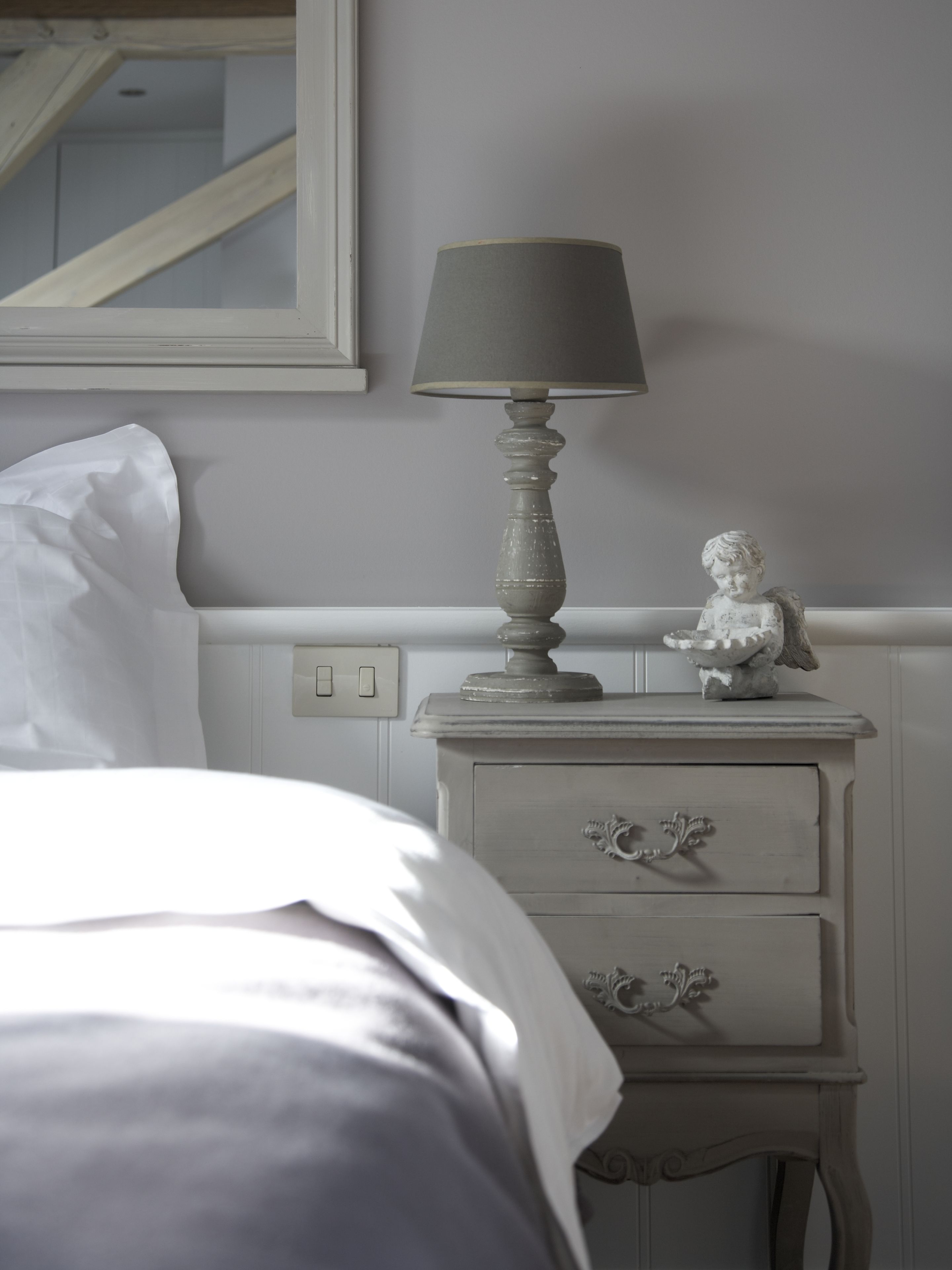 Una cama bien tendida con sábanas blancas, una mesita de noche con una lámpara y un pequeño objeto decorativo.