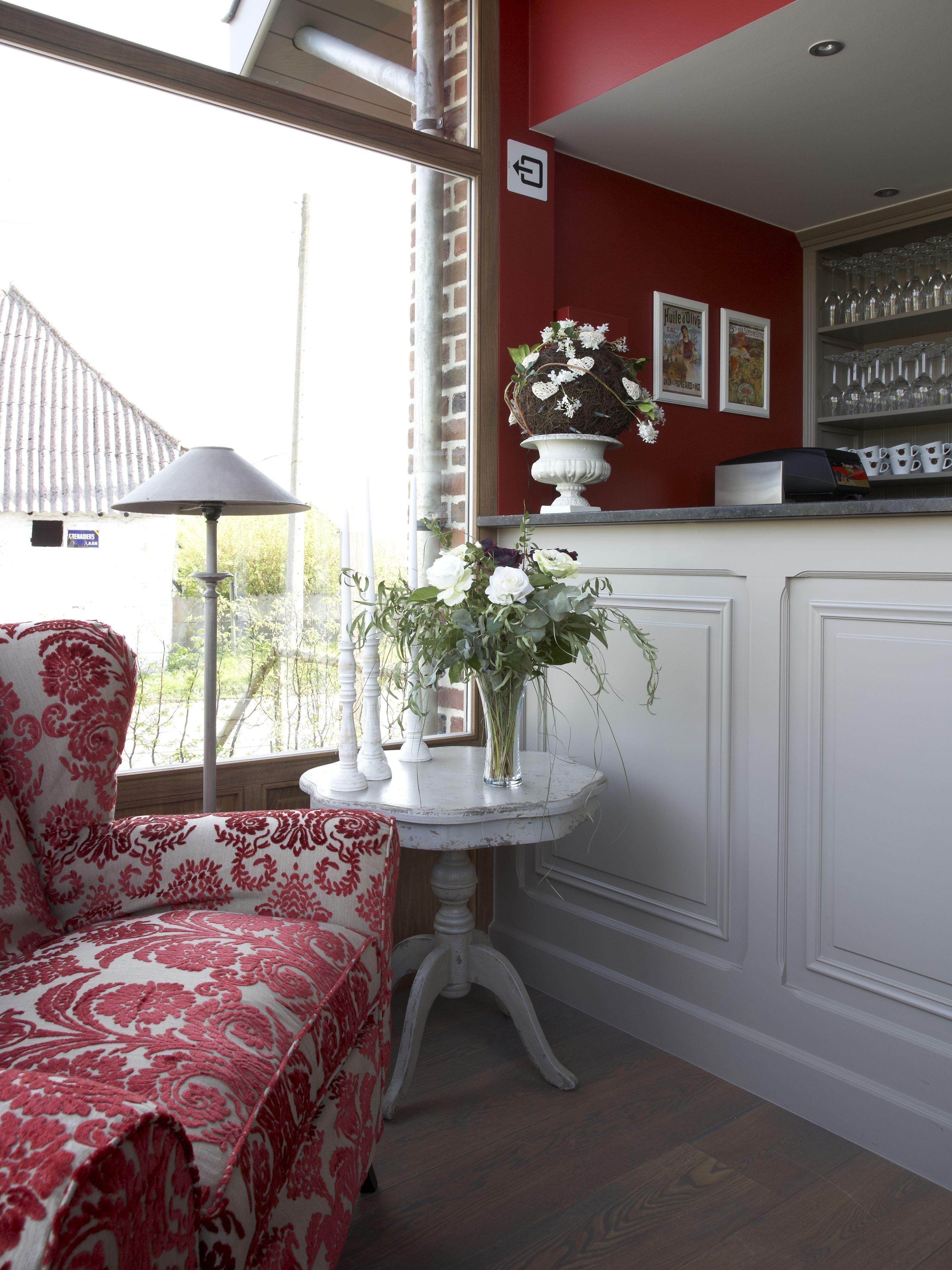 Een kamer met een rode wand, een witte stoel met een rood-wit patroon, een ronde tafel met een bloevas, en een plank met glazen en een koffieapparaat.