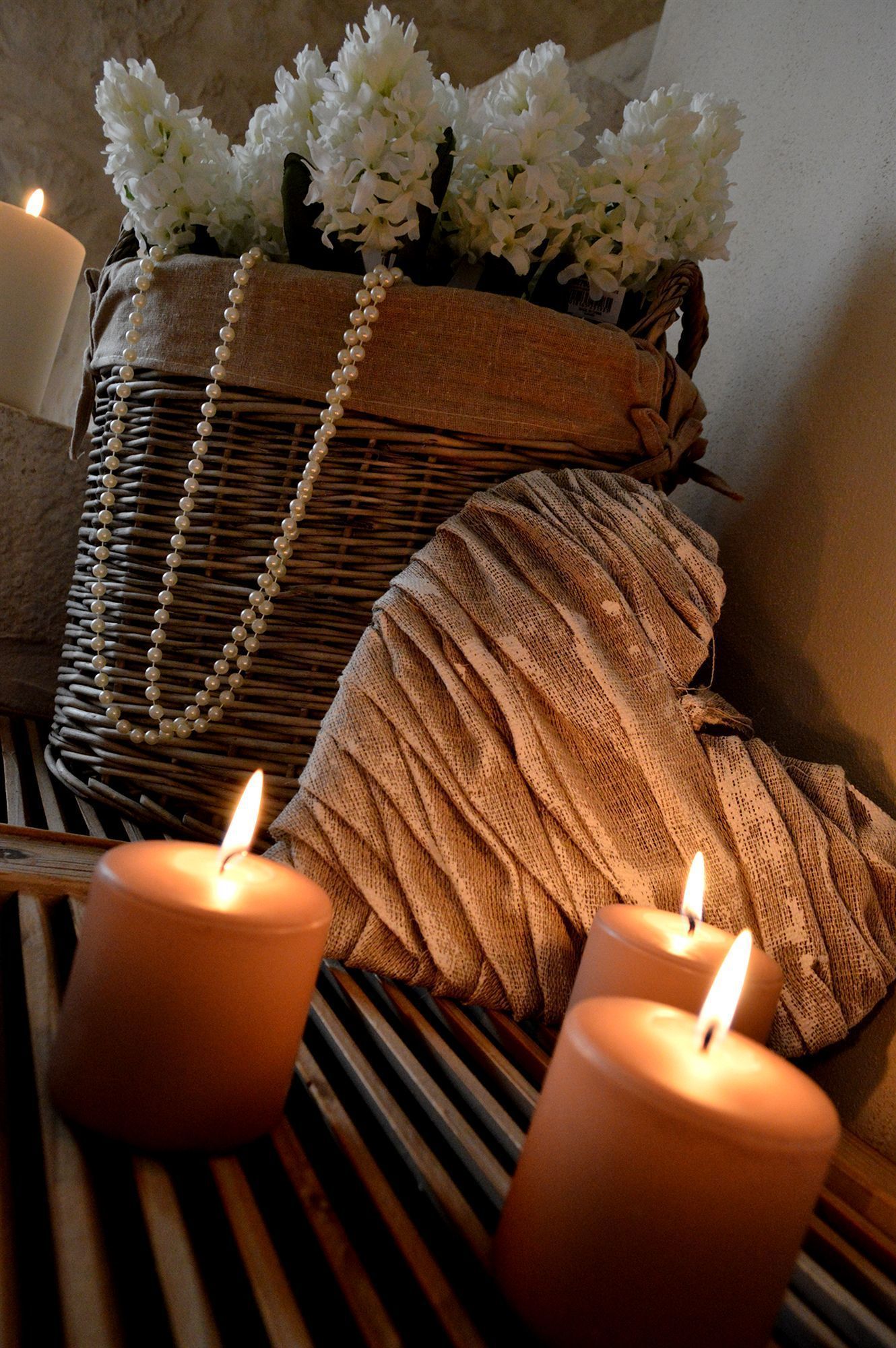 Un ambiente tranquilo con dos velas encendidas, una cesta tejida con flores blancas y una tela con textura.