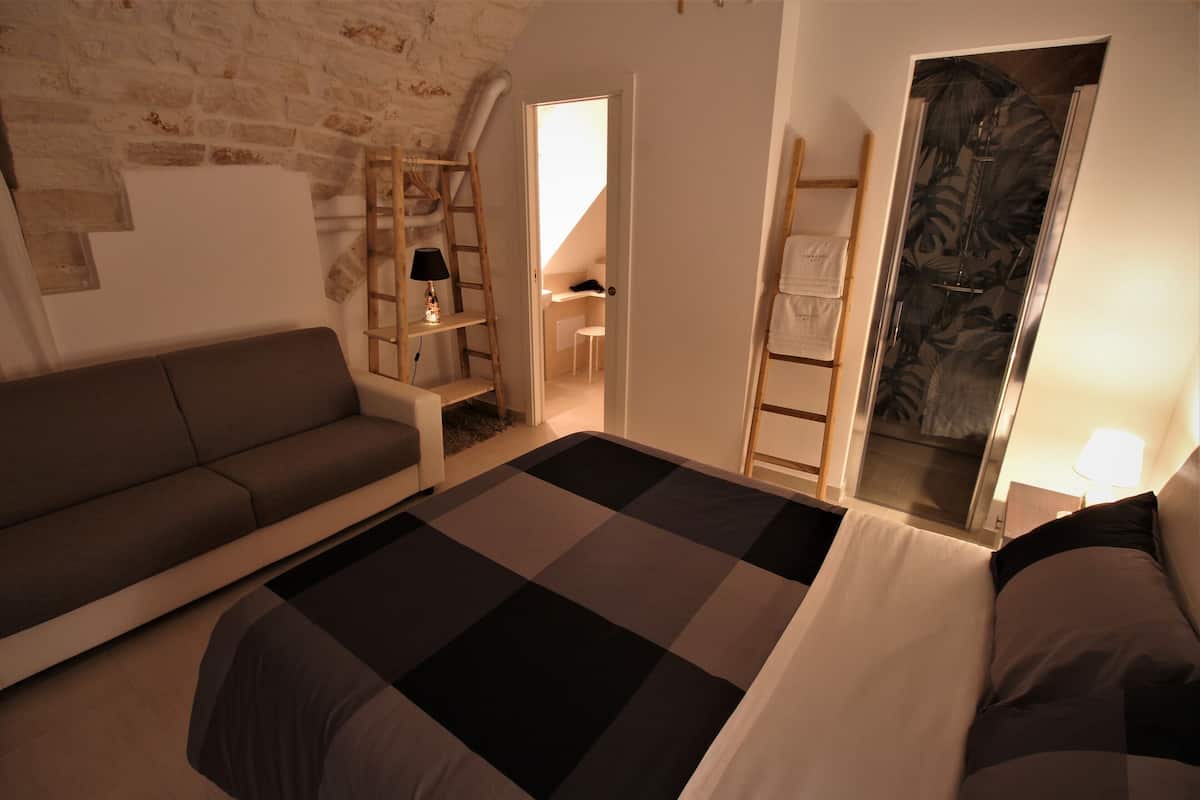 Quarto com cama, sofá, escada, escrivaninha e banheiro visível por uma porta aberta.