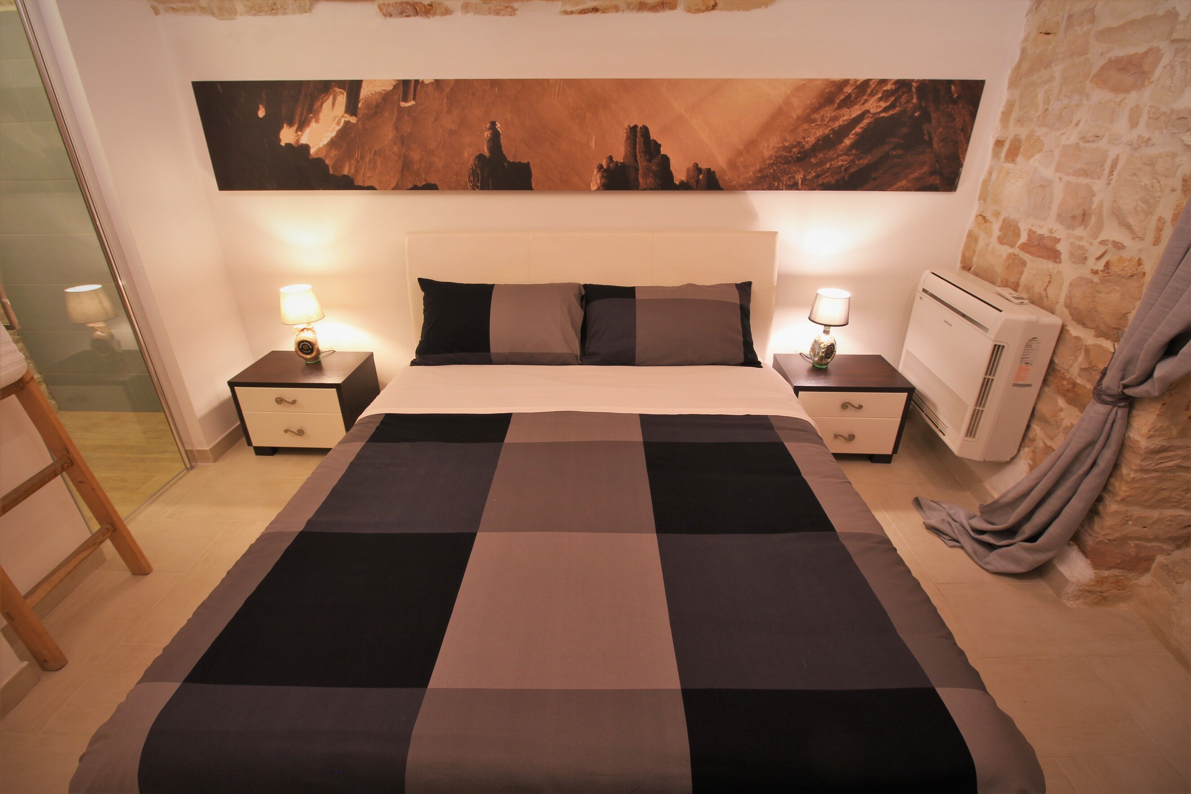 Een slaapkamer met een groot bed, twee nachtkastjes met lampen, een wandairconditioner en een stenen muur.
