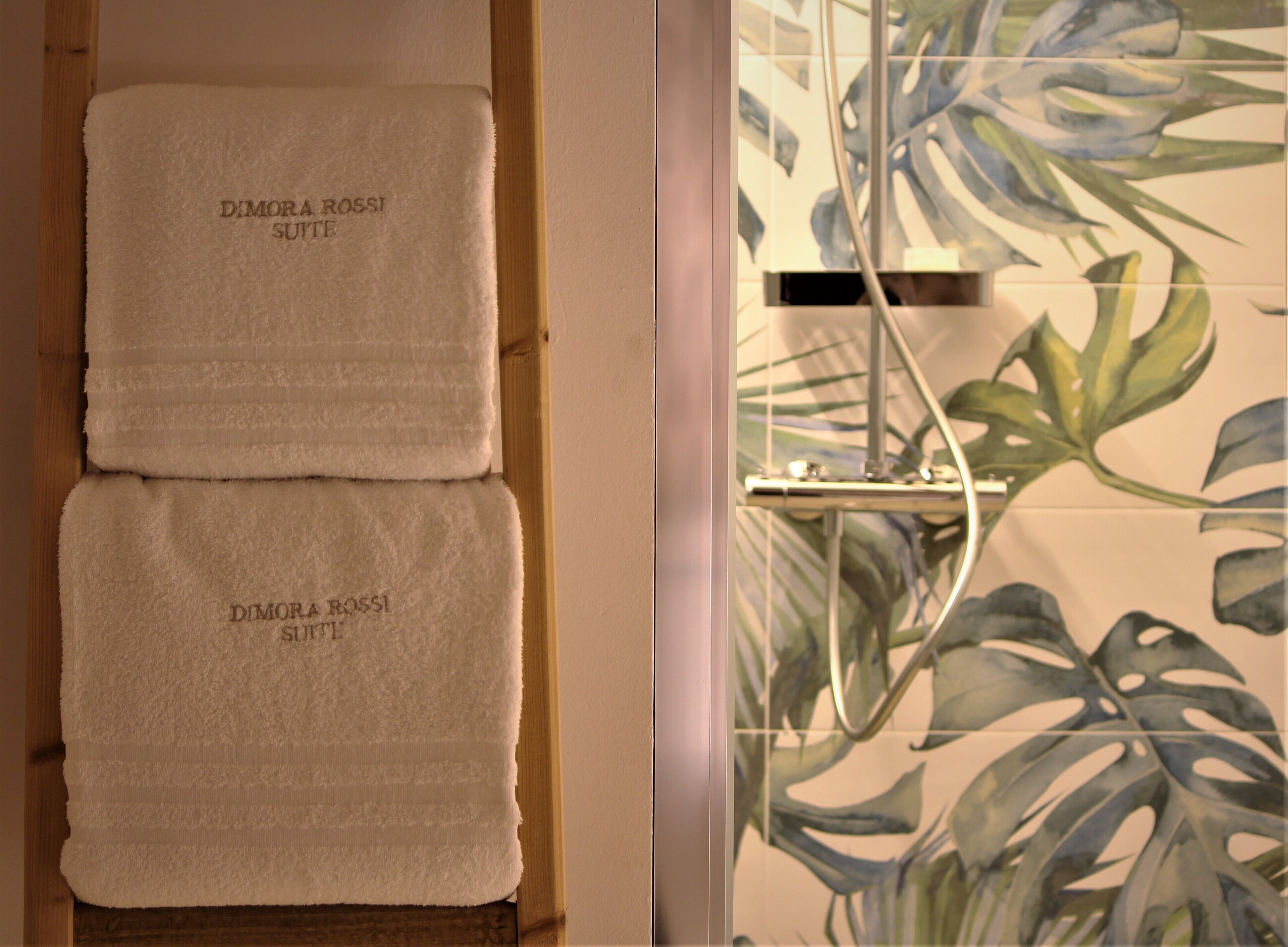 Two towels with "Dimora Rossi Suite" text, a shower with a glass door, and a plant with large leaves.