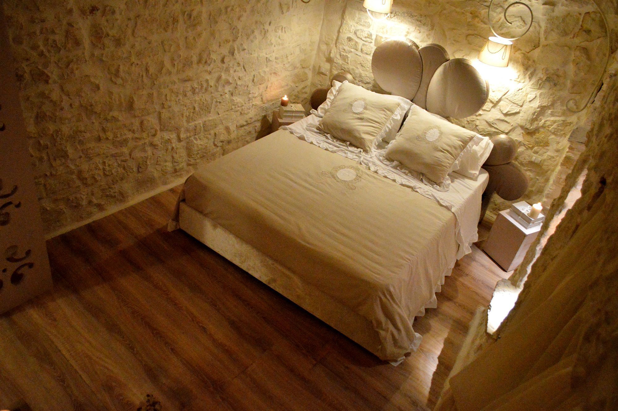 Slaapkamer met een stenen muur, houten vloer en een bed met meerdere kussens, een nachtkastje met een kaars.
