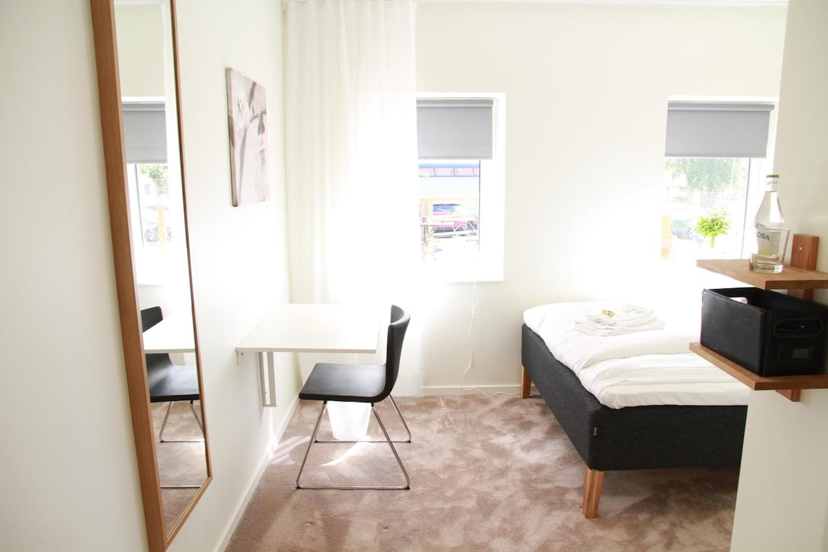 A small, modern bedroom with a bed, a desk, and a chair. There is a window with a view of the outside.