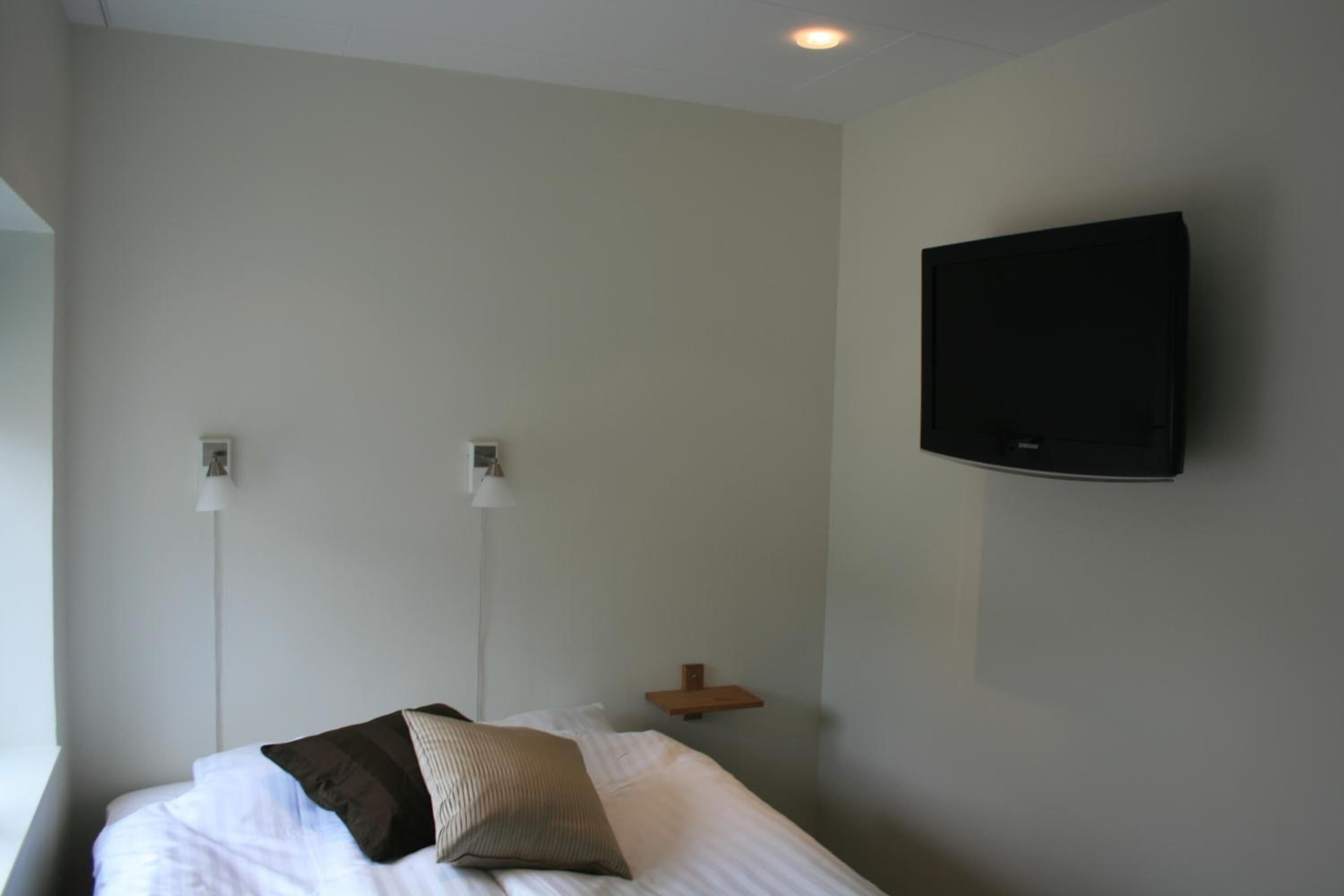 Une chambre d’hôtel avec un lit, deux lampes de chevet, un téléviseur fixé au mur et une petite étagère en bois.