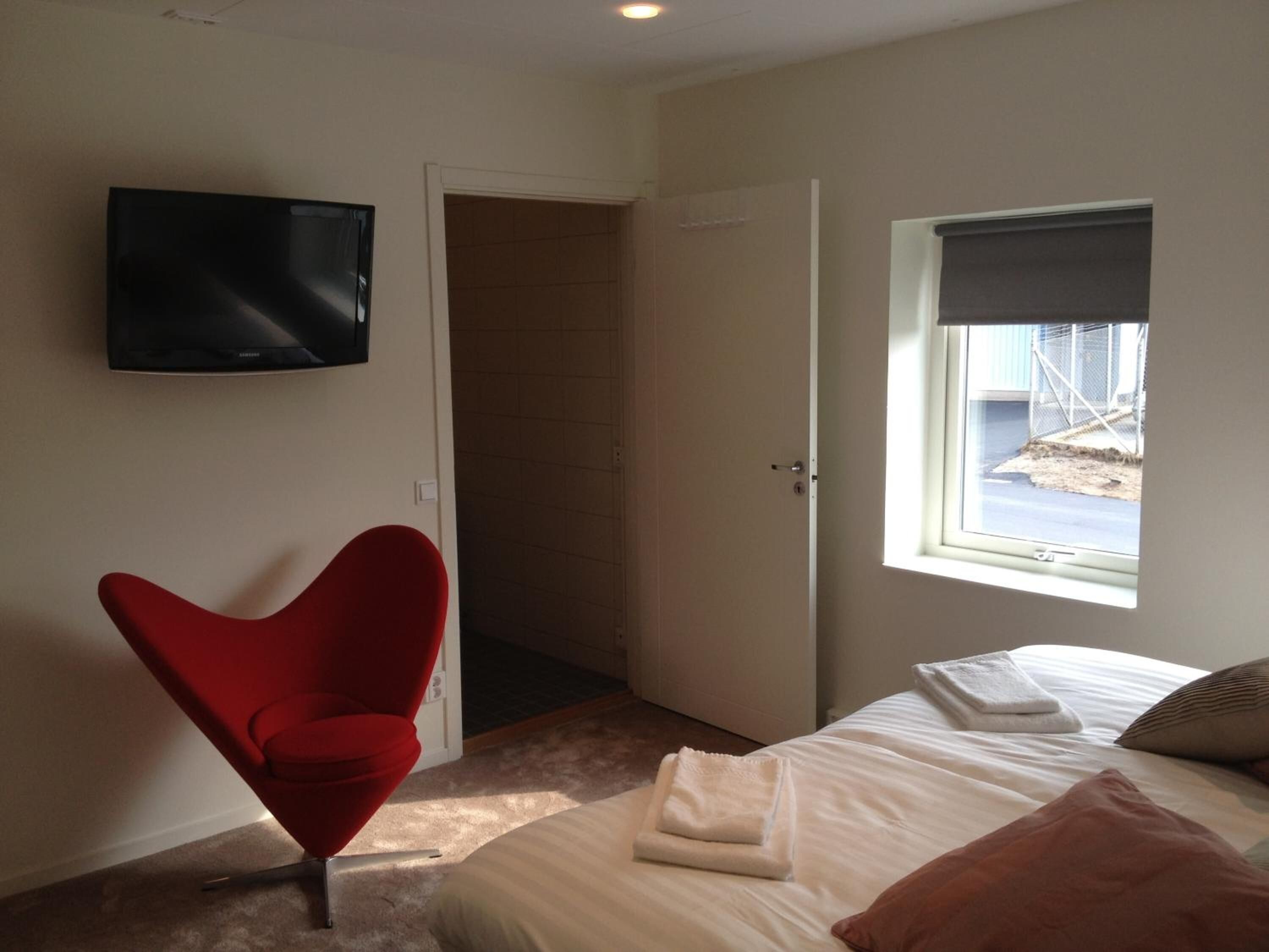 A bedroom with a bed, a red chair, a TV, and a window with a view of the outside.