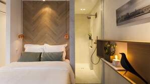 A modern hotel room with a wooden herringbone wall, a bed with white linens, a nightstand with a black bird sculpture, and a shower area with a glass door. - Kaai 11 (Antwerp)