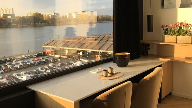 A modern hotel room with a large window overlooking a parking area and a body of water. There is a dining area with a table and chairs, and a bed with a pillow and a remote control on the side. - Kaai 11 (Antwerp)
