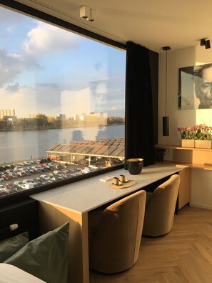 A modern hotel room with a large window overlooking a parking area and a body of water. There is a dining area with a table and chairs, and a bed with a pillow and a remote control on the side. - Kaai 11 (Antwerp)