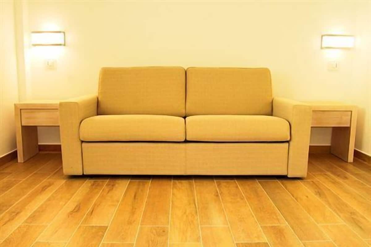 Un sofá de tonos neutros con dos mesitas auxiliares, suelo de madera y luces fijadas a la pared.