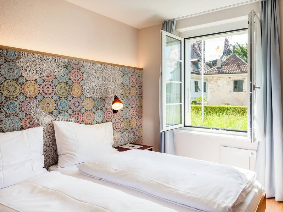 A bedroom with a bed, a nightstand, a wall-mounted lamp, and a window with curtains.