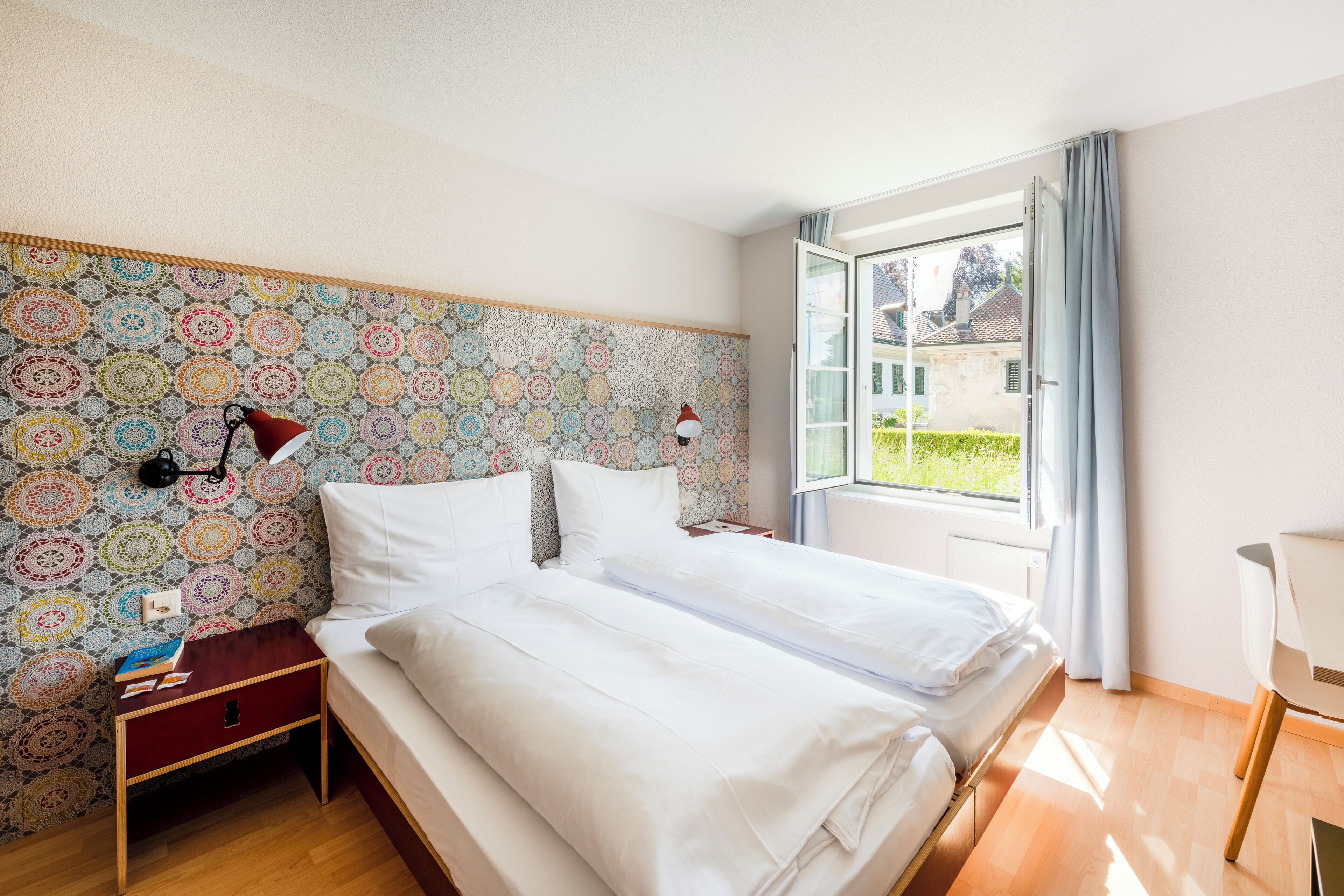 A bedroom with two beds, a patterned wallpaper, a nightstand, a lamp, and a window with curtains.