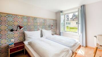 A bedroom with two beds, a patterned wallpaper, a nightstand, a lamp, and a window with curtains.