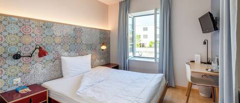 Una habitación de hotel con cama, escritorio, televisor y ventana con cortinas.