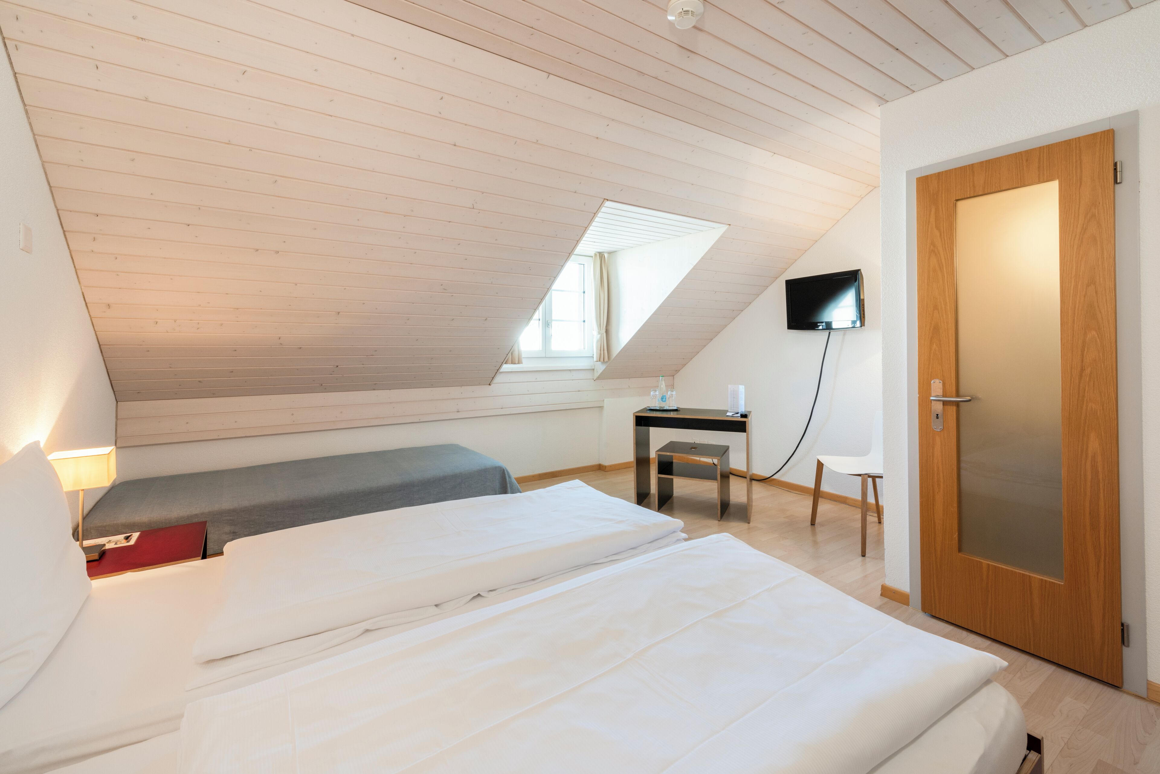 A bedroom with a sloped ceiling, a bed, a desk, a chair, a TV, and a door.