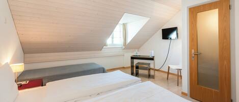 A bedroom with a sloped ceiling, a bed, a desk, a chair, a TV, and a door.