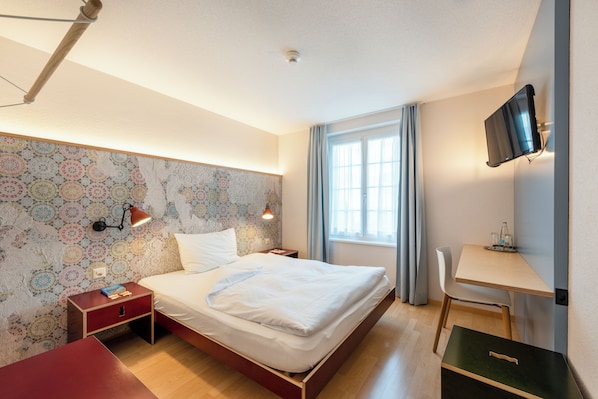 A neatly arranged bedroom with a bed, bedside table, lamp, and a desk with a chair.