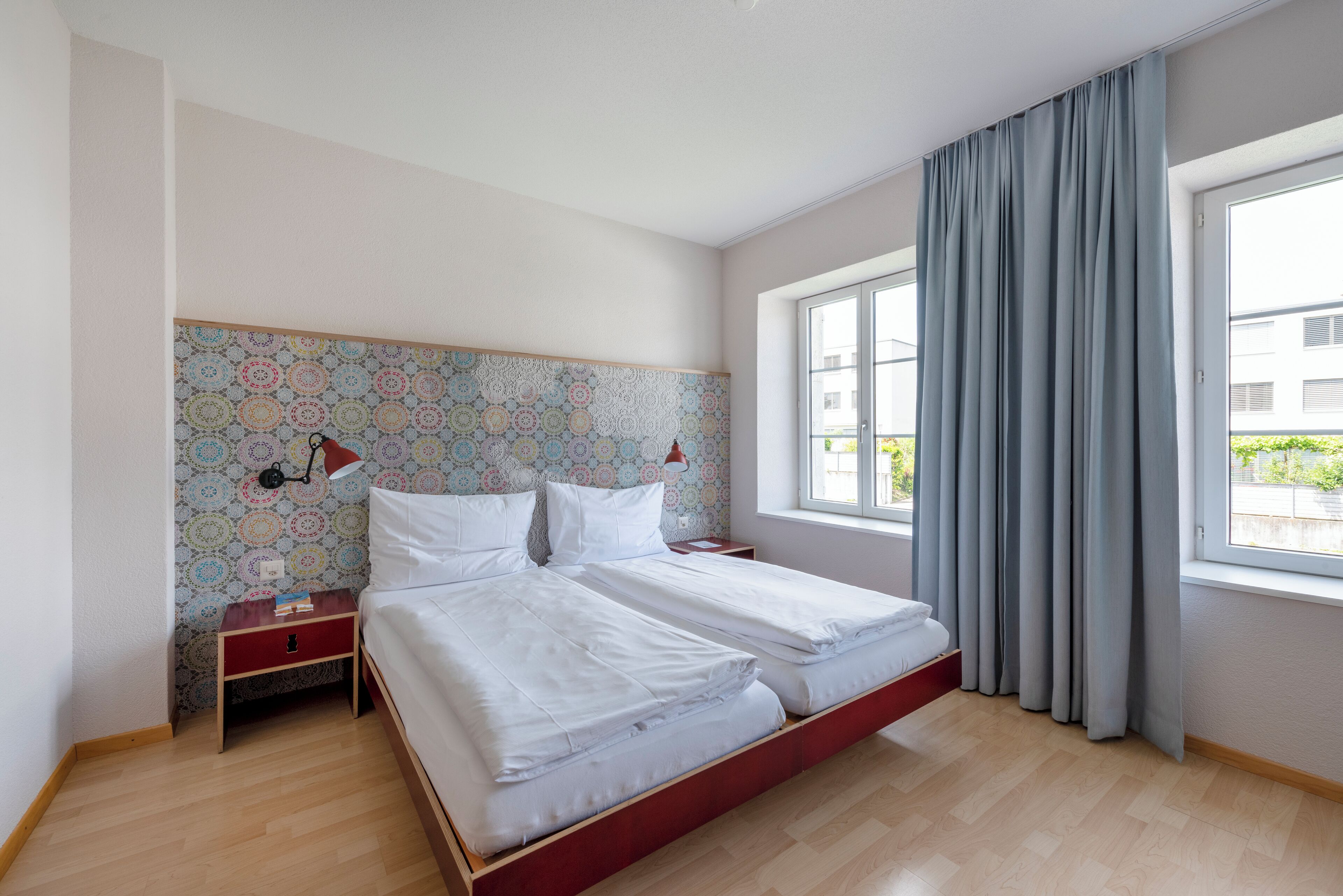 A modern bedroom with a large bed, a nightstand, a patterned wallpaper headboard, and a window with curtains.
