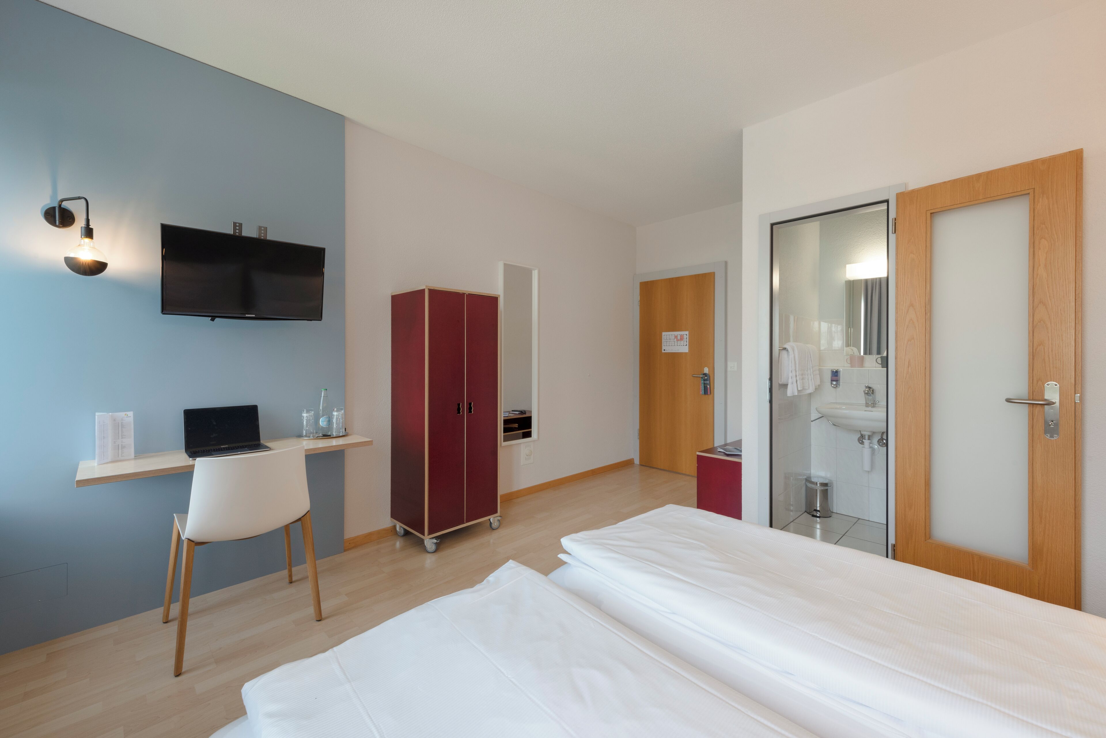 Habitación de hotel con cama, escritorio con computadora portátil, armario y baño con espejo y lavamanos.