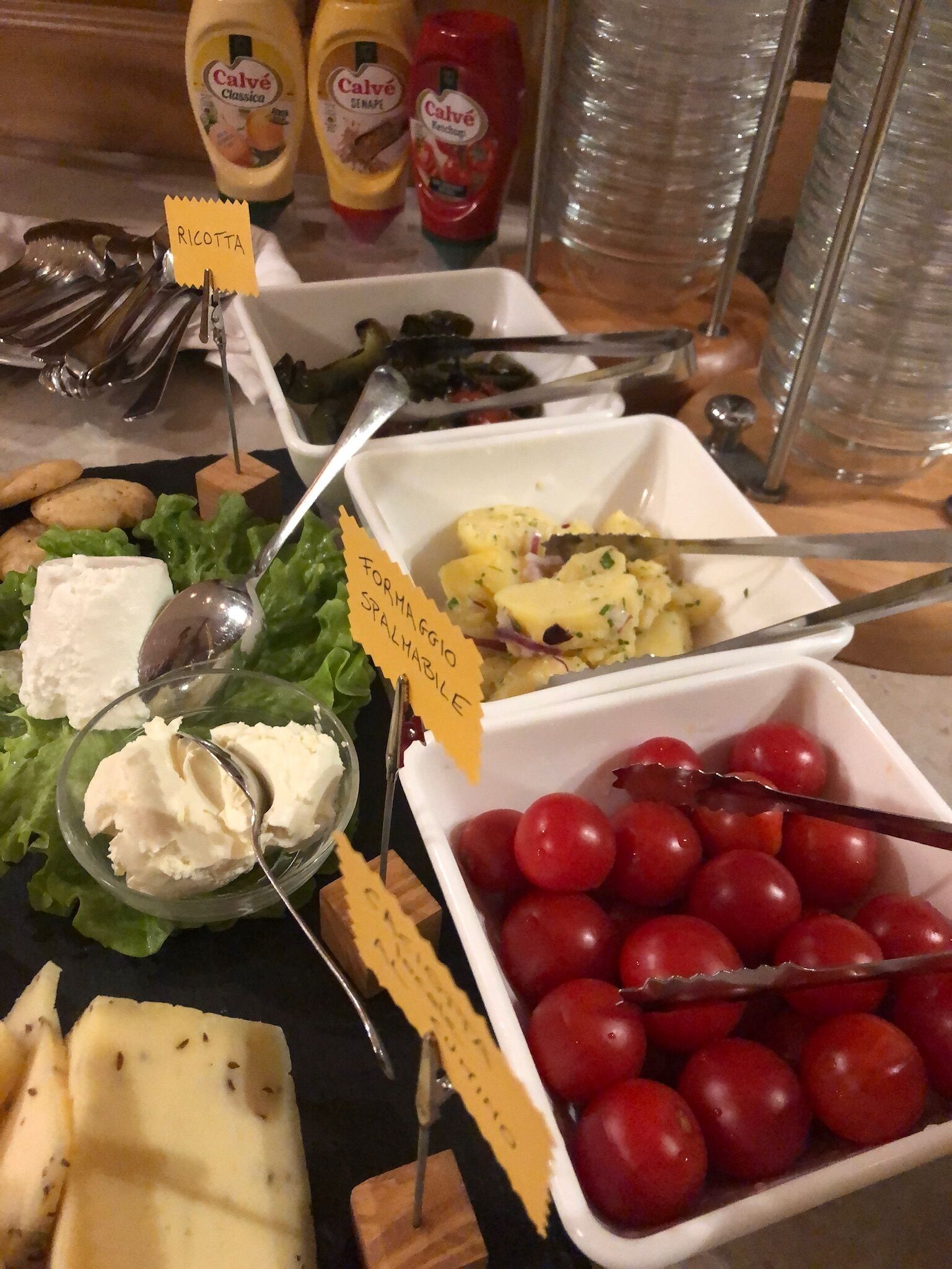 Ein reichhaltiges Buffet mit verschiedenen Speisen, darunter Käse, Kirschtomaten und Salat, mit Schildern, die die Art der Speisen kennzeichnen.