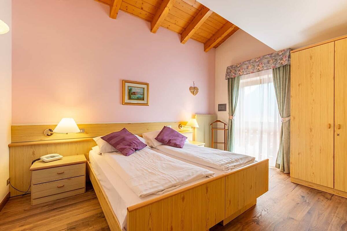 A double bed with two pillows, a bedside table with a lamp, a wooden wardrobe, and a window with curtains.