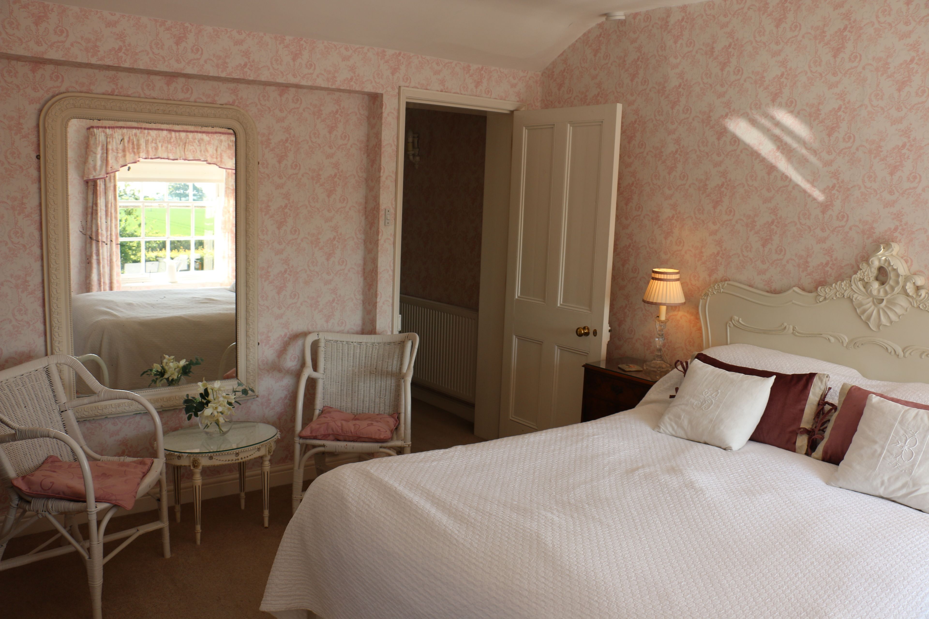 Une chambre à coucher comprenant un lit, deux chaises, une petite table avec des fleurs, un miroir et une porte donnant sur une autre pièce.