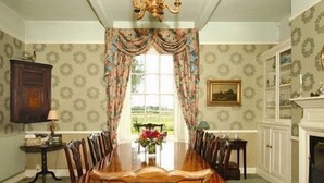 A dining room with a long wooden table, chairs, a chandelier, patterned wallpaper, a window with curtains, a fireplace, and a framed picture on the wall. - The Grange (Market Rasen)