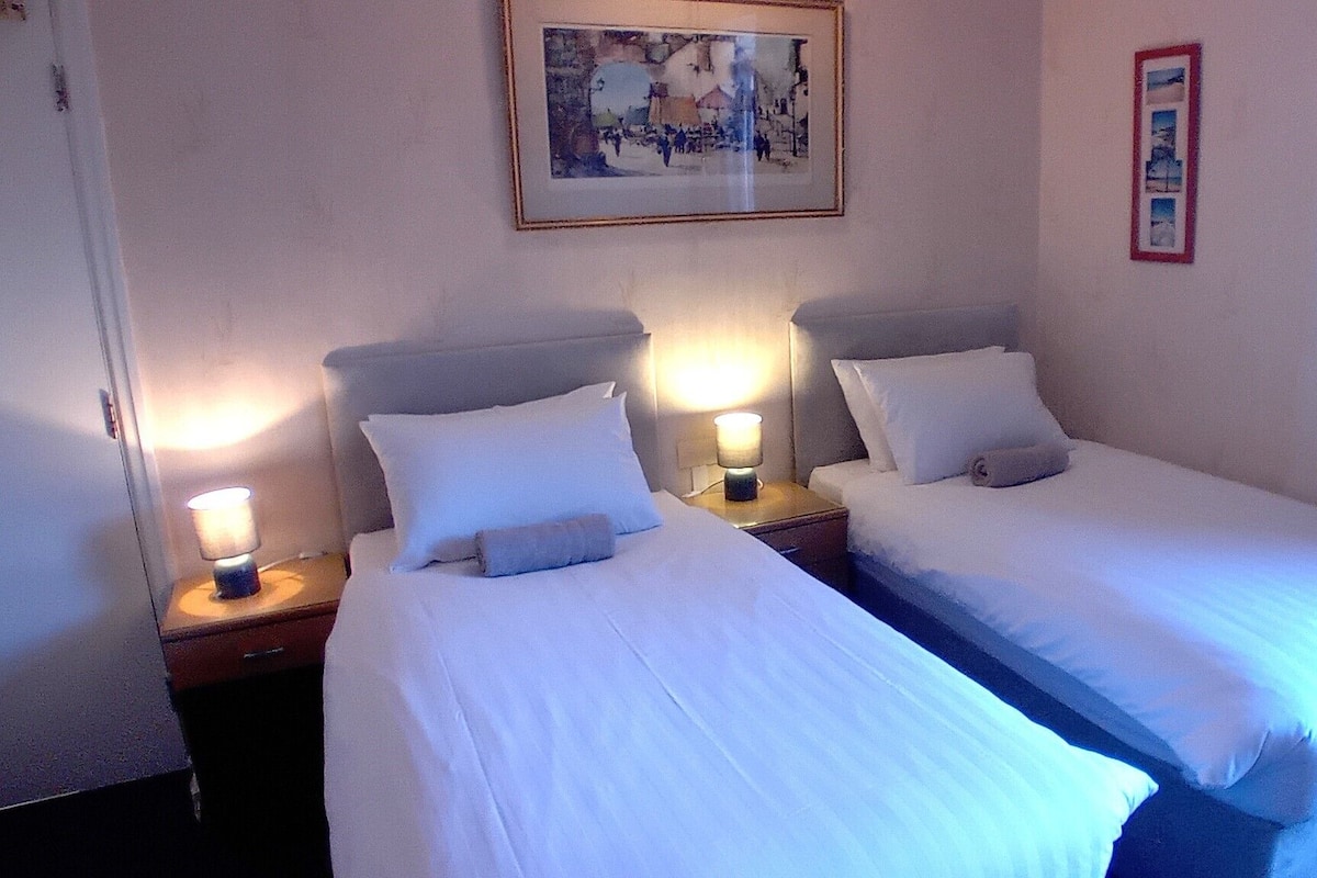 Una habitación de hotel con dos camas individuales, lámparas de noche, un cuadro enmarcado en la pared y una ventana con cortinas.