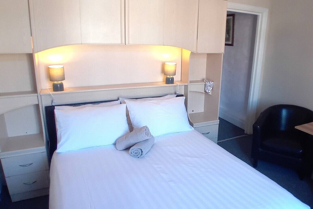 Una habitación de hotel con cama, dos lámparas de noche, una silla, una mesita y una puerta.