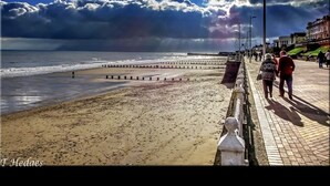 A beach with a paved walkway, people walking, and a Ferris wheel in the background. - Home and Away Guesthouse  (Bridlington)