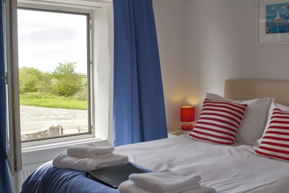 A hotel room with a bed, white bedding, red and white striped pillows, a window with blue curtains, and a view of greenery outside.
