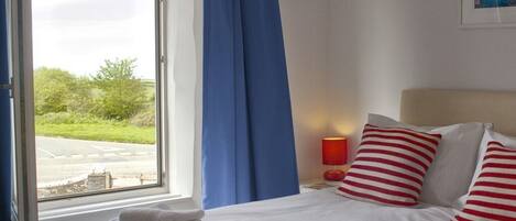 Habitación de hotel con cama, ropa de cama blanca, almohadas a rayas rojas y blancas, una ventana con cortinas azules y vistas a la vegetación.