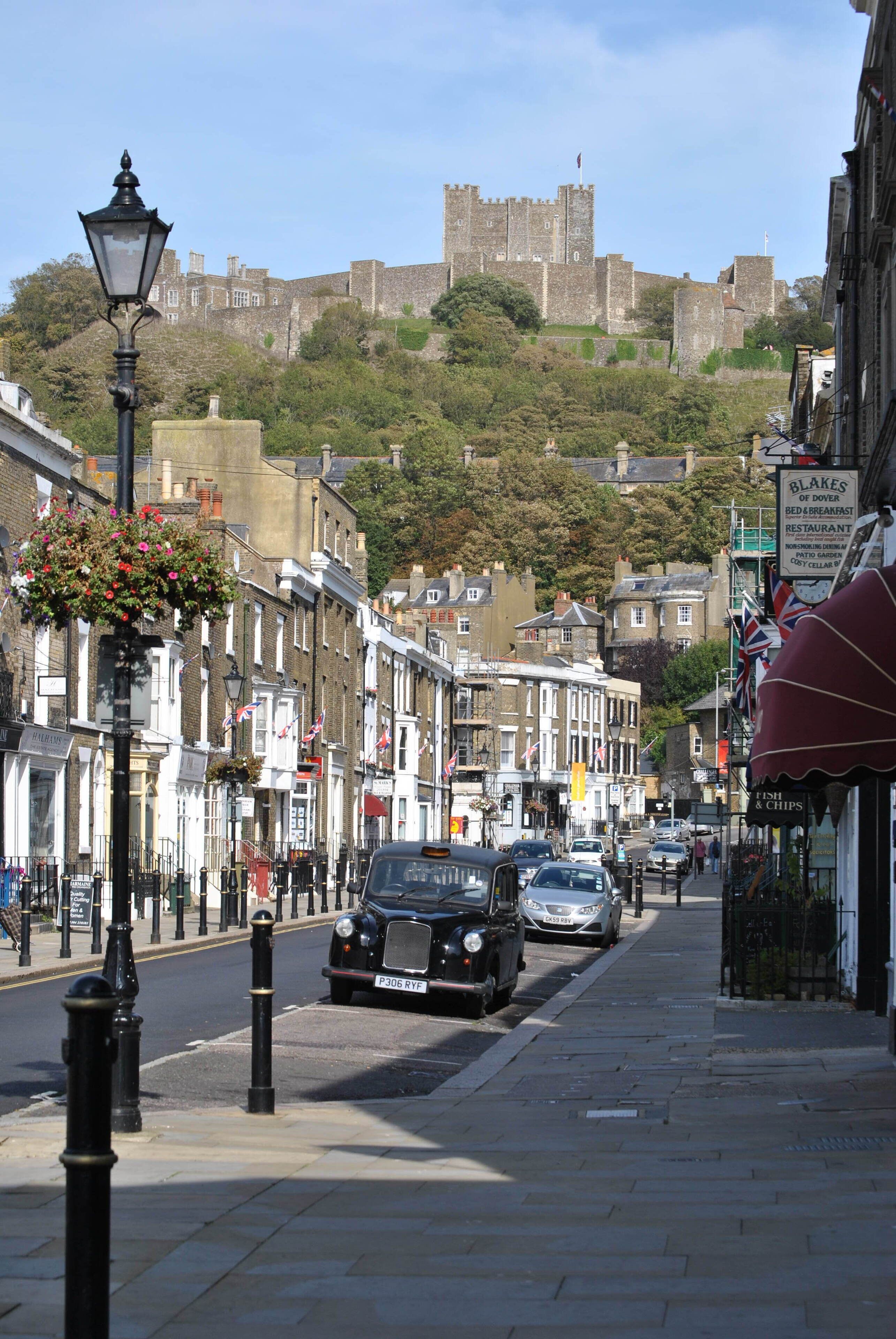Una calle con edificios históricos, un castillo imponente al fondo, y un coche negro estacionado al costado.