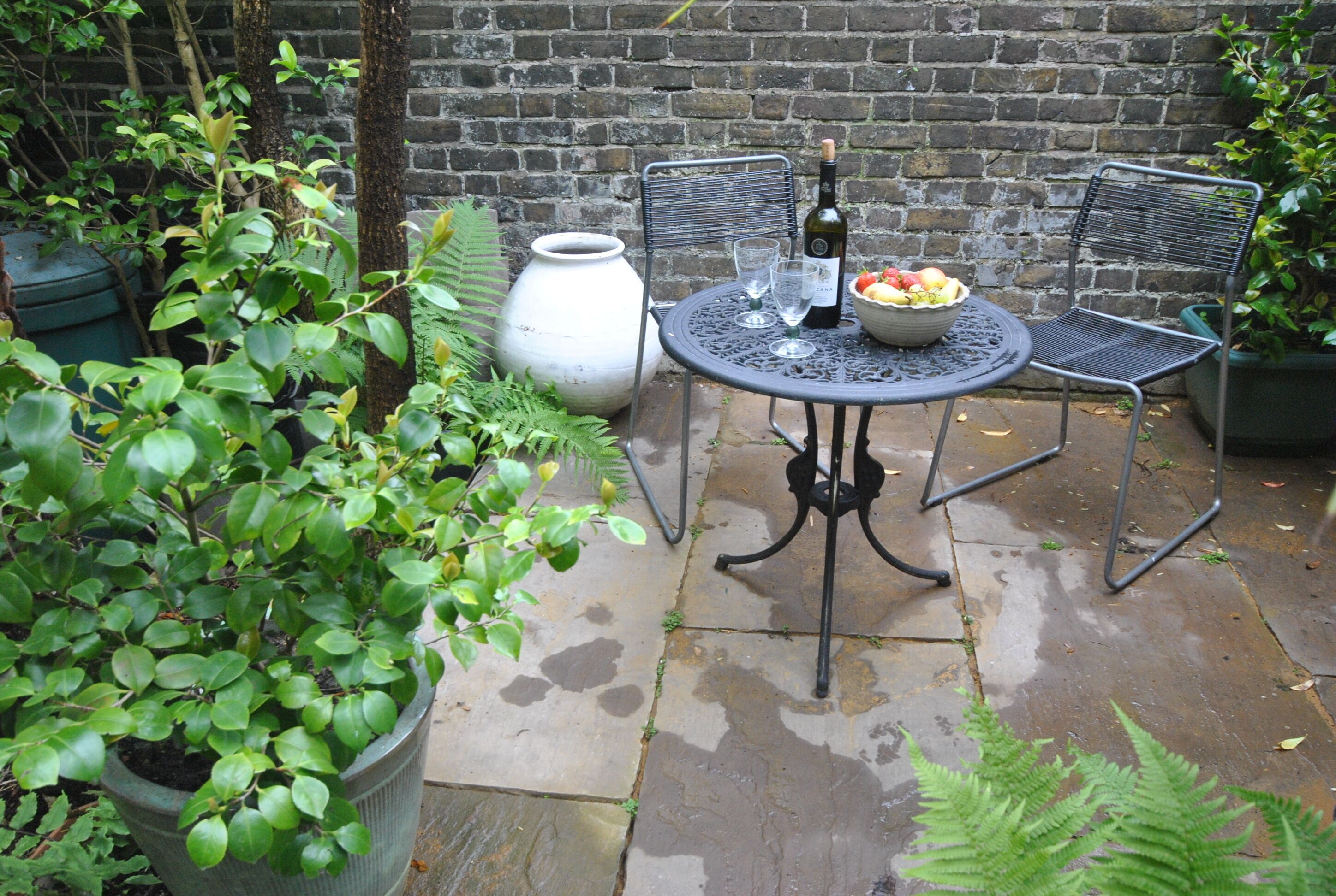 Een terras met een ronde metalen tafel, twee stoelen, een fles wijn en een schaal fruit.