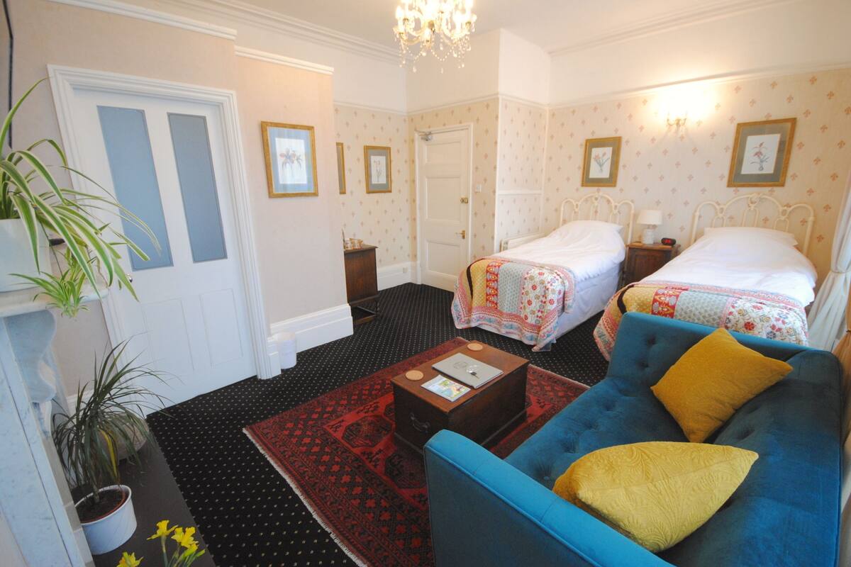 A hotel room with two beds, a blue sofa, a red patterned rug, and a potted plant.