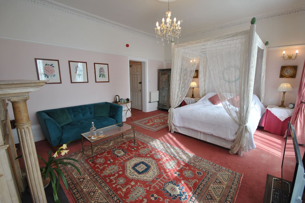 A bedroom with a four-poster bed, a sitting area with a sofa, a chandelier, and a large rug.