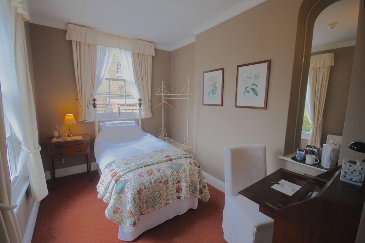 A hotel room with a bed, a desk with a chair, a window with curtains, a bedside table with a lamp, and framed pictures on the wall.