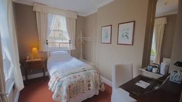 A hotel room with a bed, a desk with a chair, a window with curtains, a bedside table with a lamp, and framed pictures on the wall.